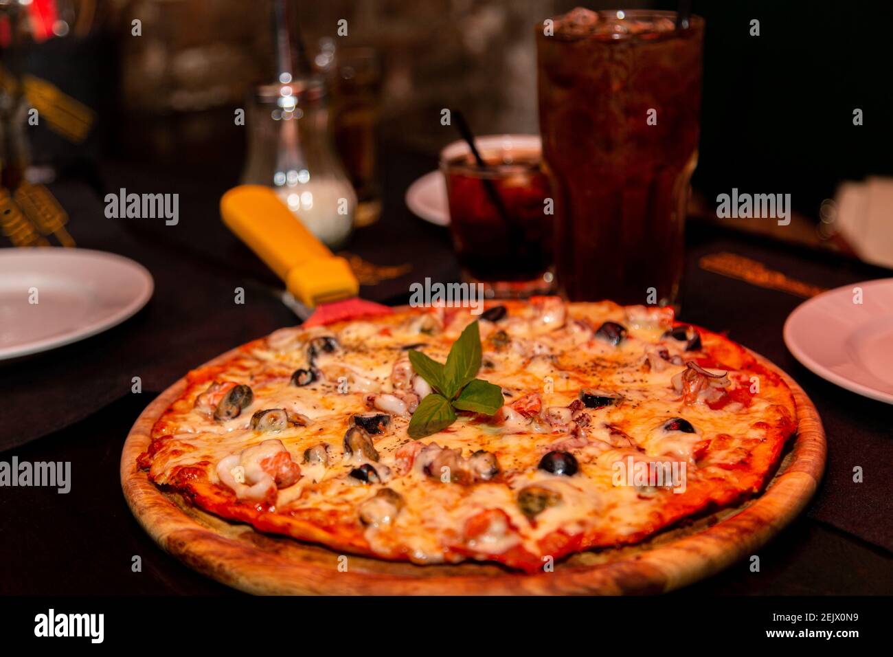 Una pizza appetitosa con frutti di mare e formaggi si trova su un piatto di legno sul tavolo. Con una bevanda fredda marrone ghiacciata. Foto Stock