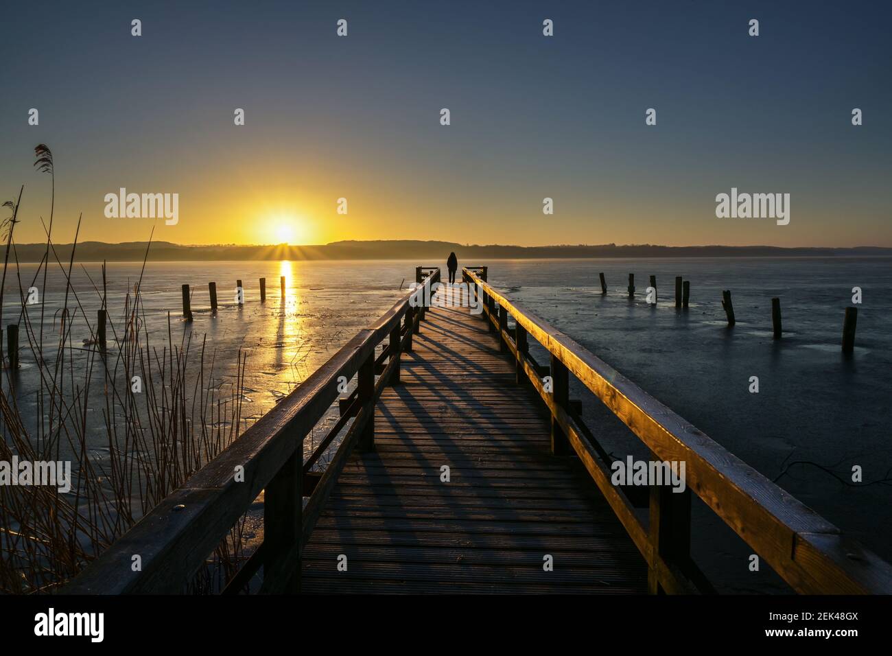 Alba su un lago ghiacciato e una giovane donna come silhouette alla fine di un lungo molo di legno godendo il paesaggio, spazio copia, selezionato focu Foto Stock