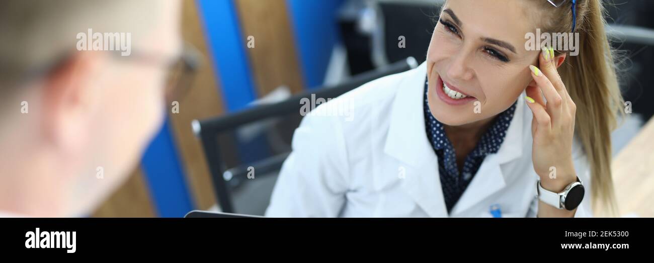 La lavorante femminile si siede sulla sedia, esamina il biomateriale al microscopio e comunica con il pacient in clinica. Foto Stock