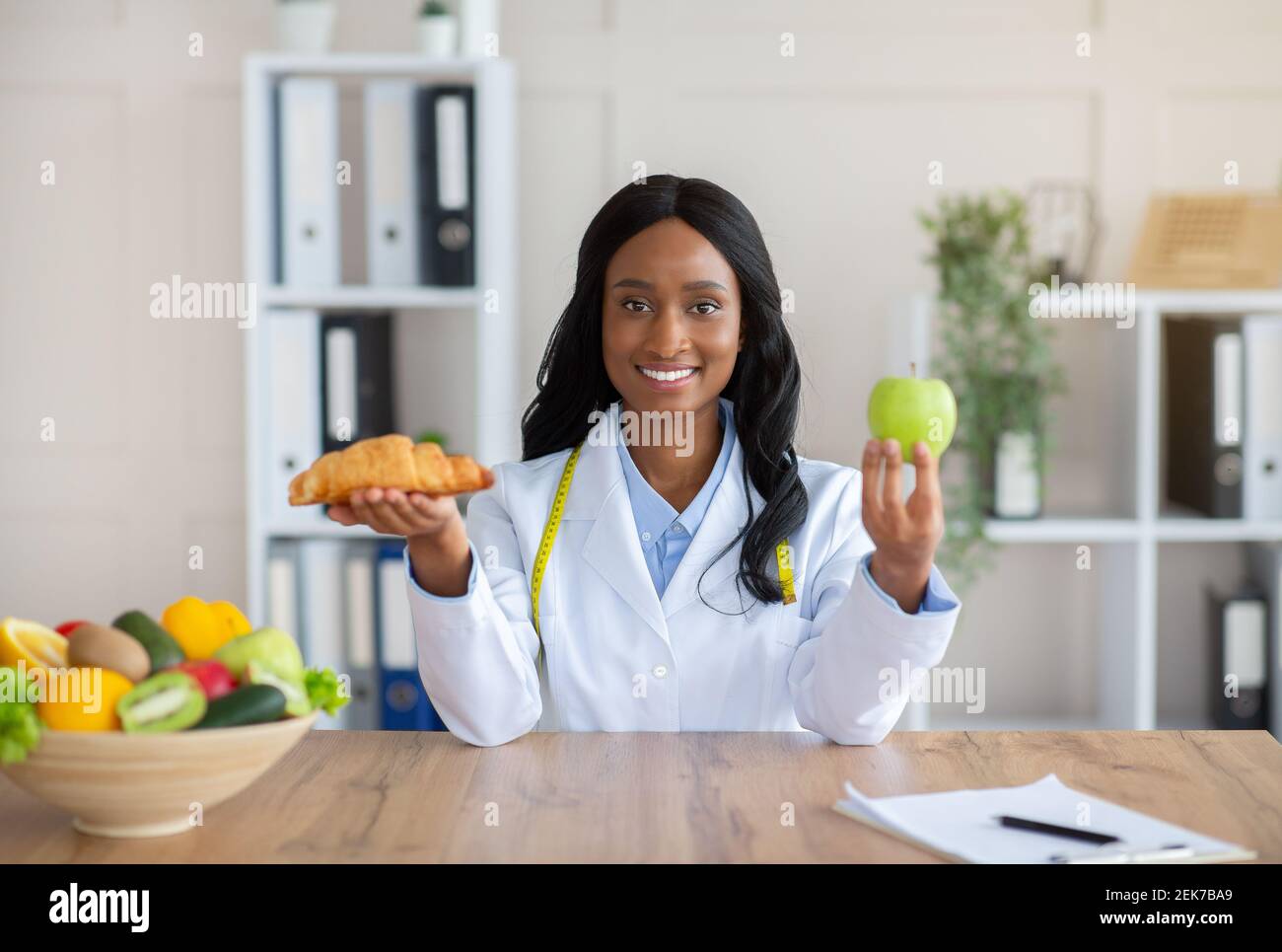 Fare scelte alimentari sane. Bella nutrizionista femminile che tiene croissant e mela al tavolo in ufficio medico Foto Stock