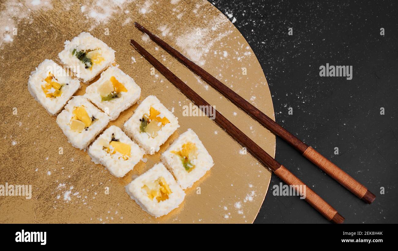 Consegna sushi. Panini dolci a base di riso, ananas, kiwi e mango. Rotola su sfondo oro e nero. Bastoncini di legno per sushi. Foto Stock