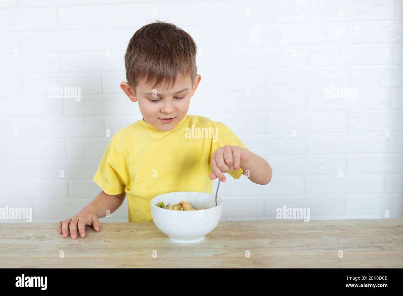 Ragazzo mancino in una T-shirt gialla mangiando insalata di frutta e sorridendo. Concetto di cibo sano per i bambini. Prodotti nutrizionali. Assistenza sanitaria Foto Stock