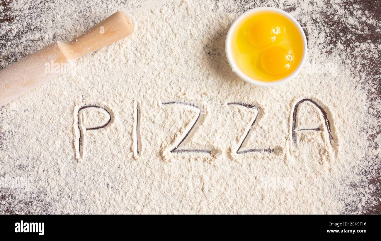 L'iscrizione Pizza su farina spruzzata, disegnata su fondo di legno, spilla e uova. Ricette di cottura. Foto Stock