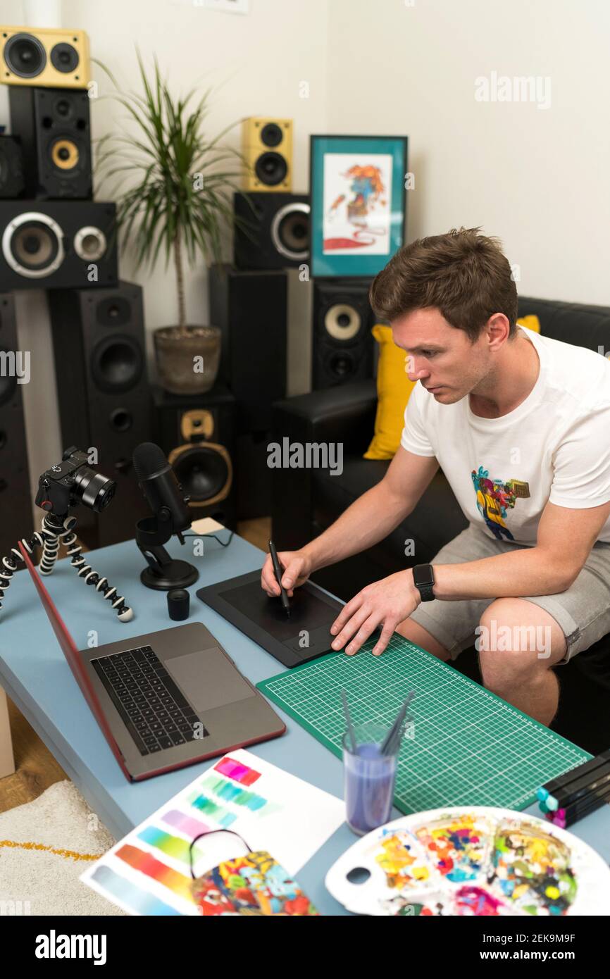 Artista maschile che utilizza il pad di schizzo durante lo streaming live attraverso la fotocamera seduto a casa Foto Stock
