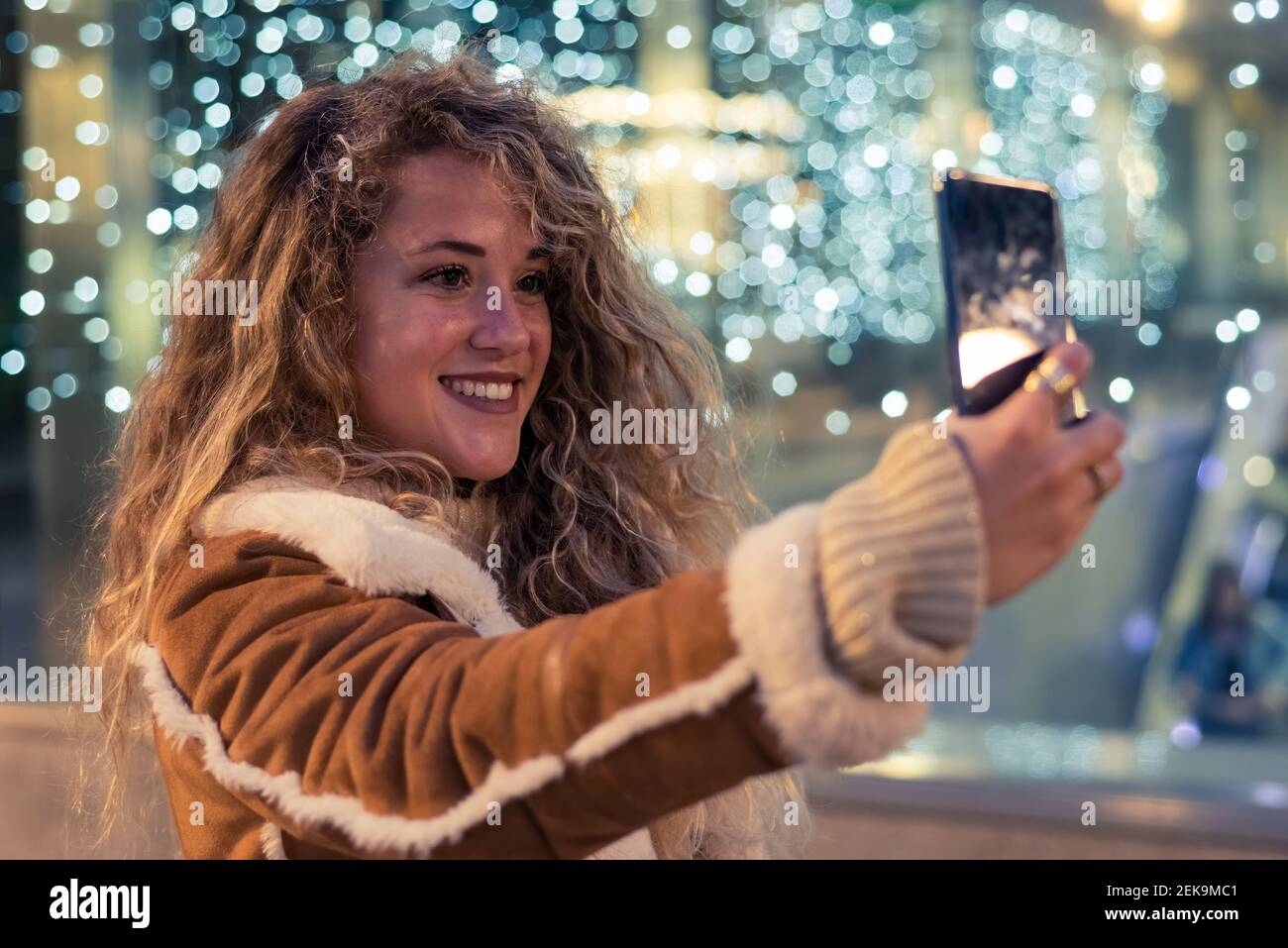 Giovane donna con capelli crespi che prende selfie attraverso smartphone contro parete di vetro illuminata Foto Stock