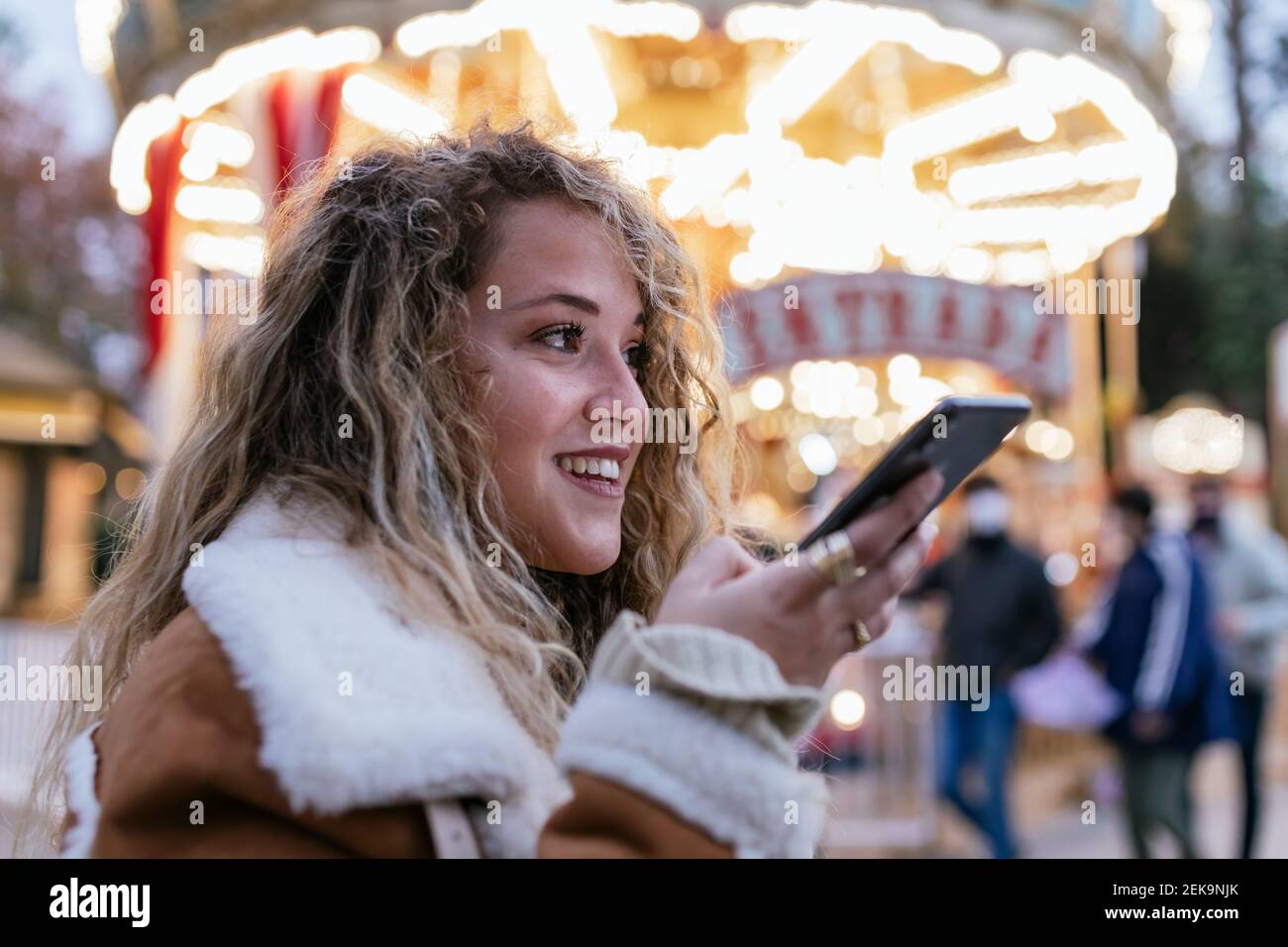 Giovane donna che invia messaggi vocali tramite smartphone mentre si è in piedi contro la giostra illuminata Foto Stock