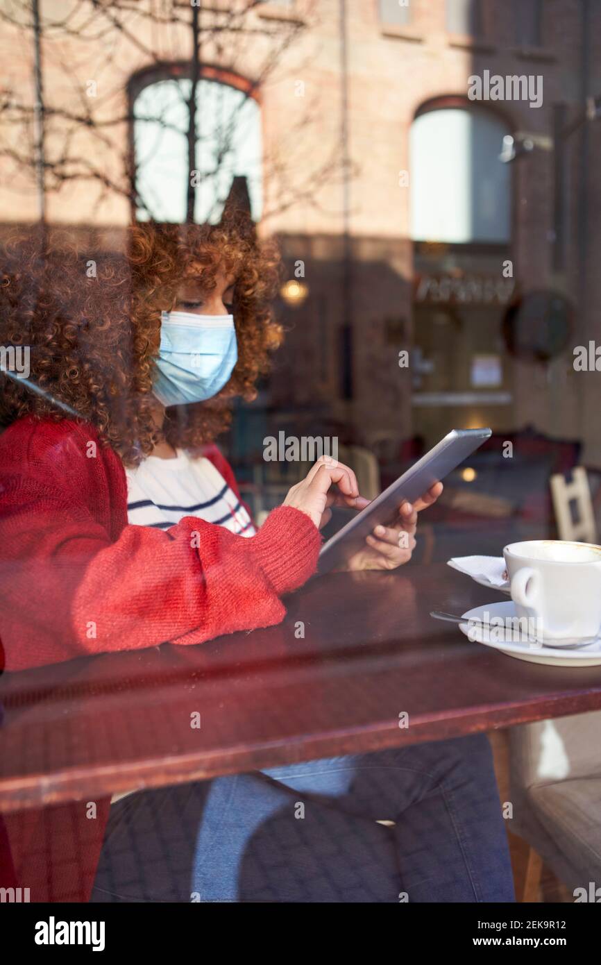 Giovane donna che indossa una maschera protettiva per il viso utilizzando un tablet digitale mentre seduto vicino alla finestra al caffè Foto Stock