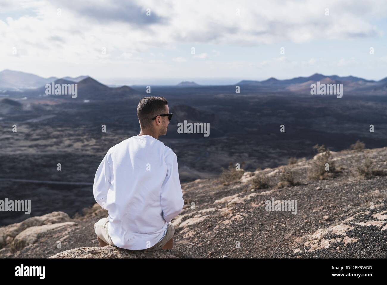 Turista maschile seduto sulla roccia mentre si guarda il vulcano El Cuervo in giornata di sole, Lanzarote, Spagna Foto Stock