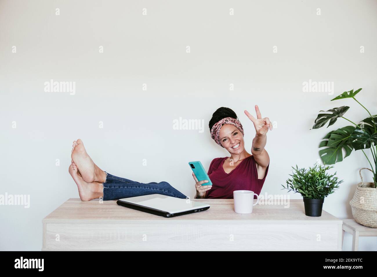 Allegro imprenditore femminile che fa segno di pace mentre si rilassa con i piedi in ufficio a casa Foto Stock