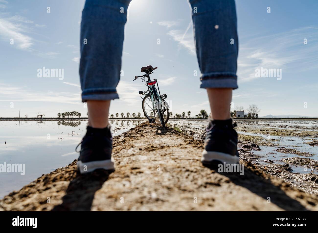 Bicicletta vista attraverso gambe di donna in piedi sul sentiero del Delta di Ebro durante il giorno di sole, Spagna Foto Stock
