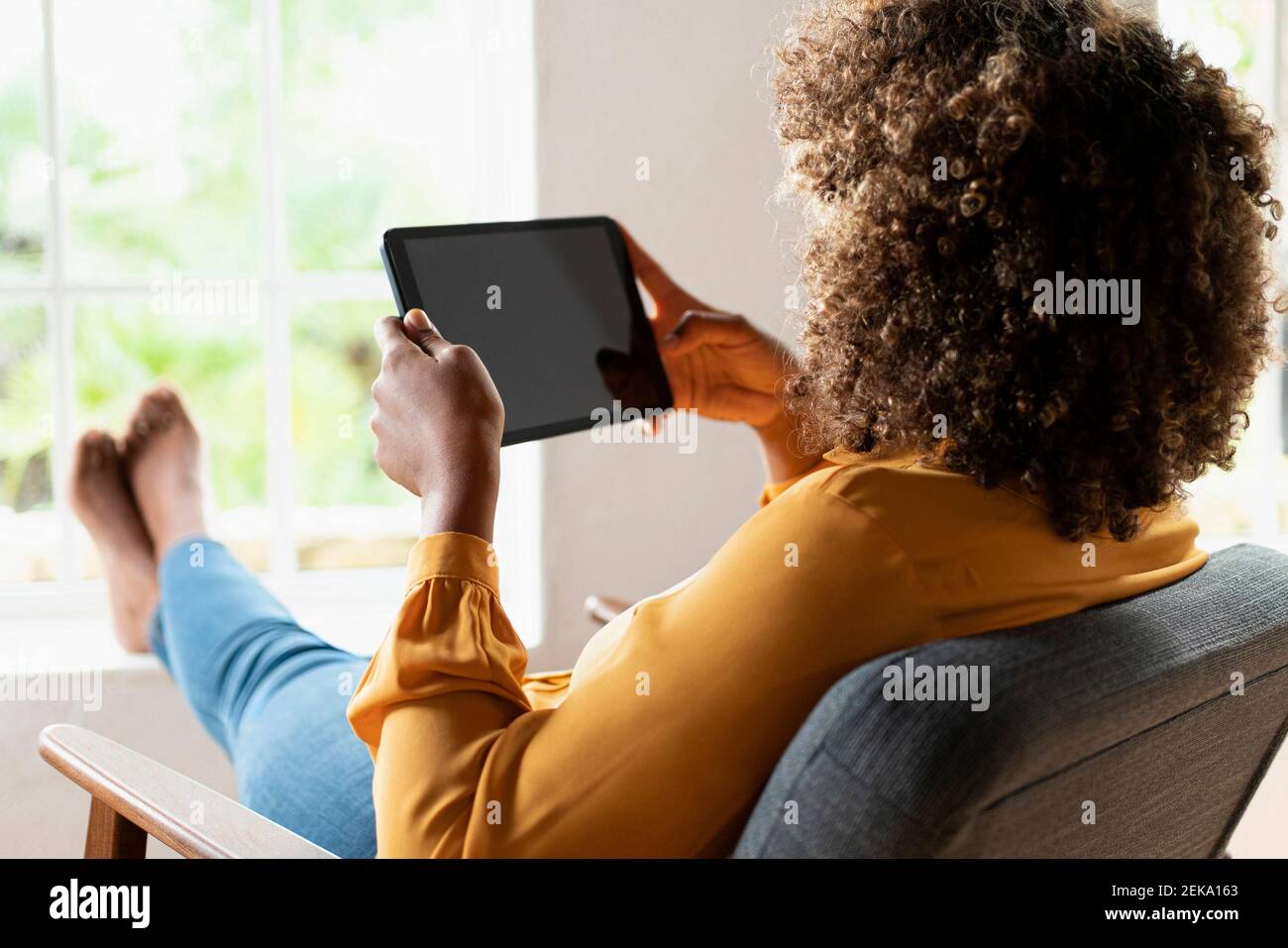 Donna con capelli ricci che usa il tablet digitale mentre si riposa sulla poltrona a casa Foto Stock