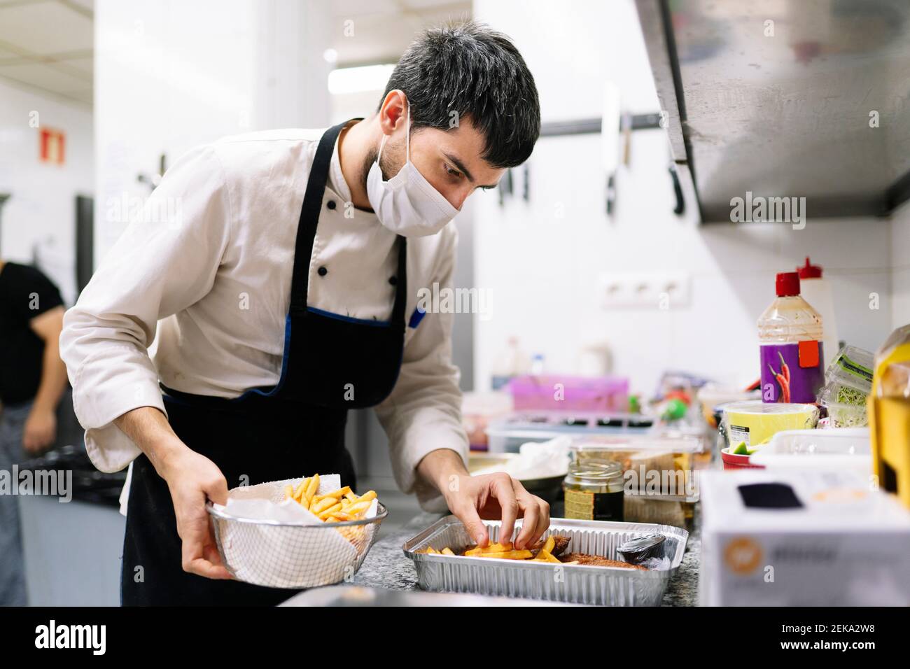 Chef maschile in maschera protettiva che dispone patatine fritte in contenitore in alluminio nella cucina del ristorante Foto Stock