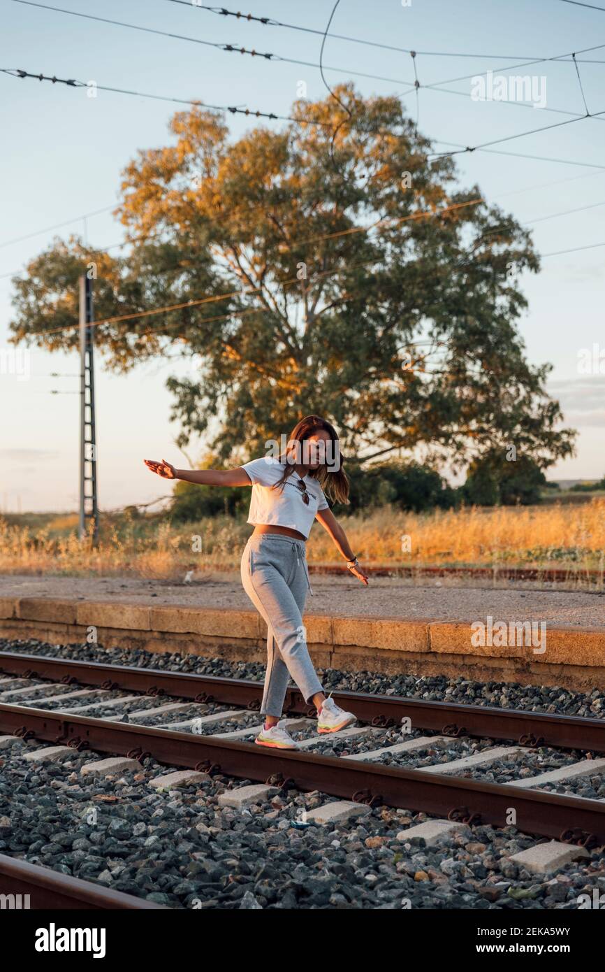 Giovane donna spensierata con le braccia allungate che camminano sulla pista ferroviaria durante il tramonto Foto Stock