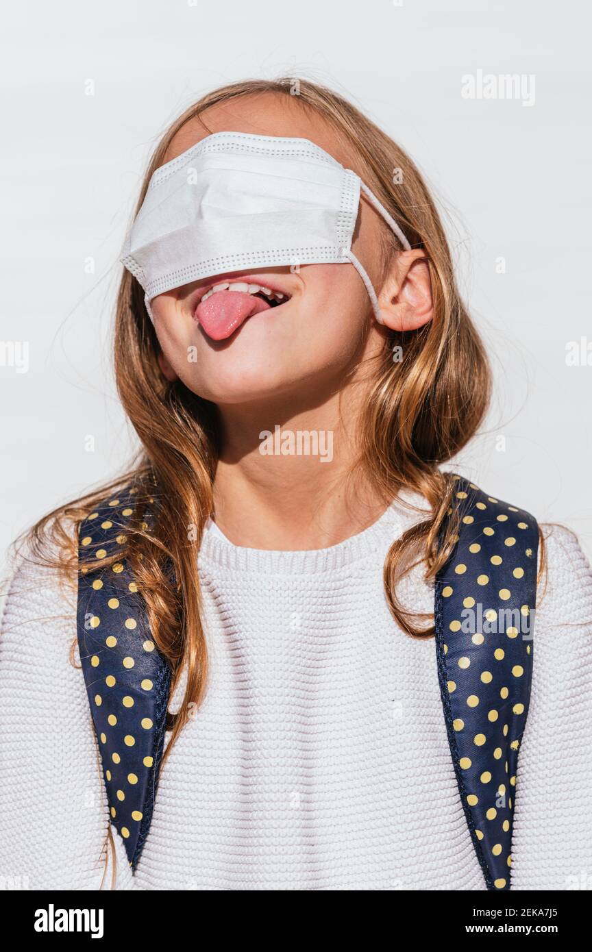 Ragazza giocosa che si stacca la lingua mentre copre gli occhi con protettivo Maschera facciale durante COVID-19 Foto Stock