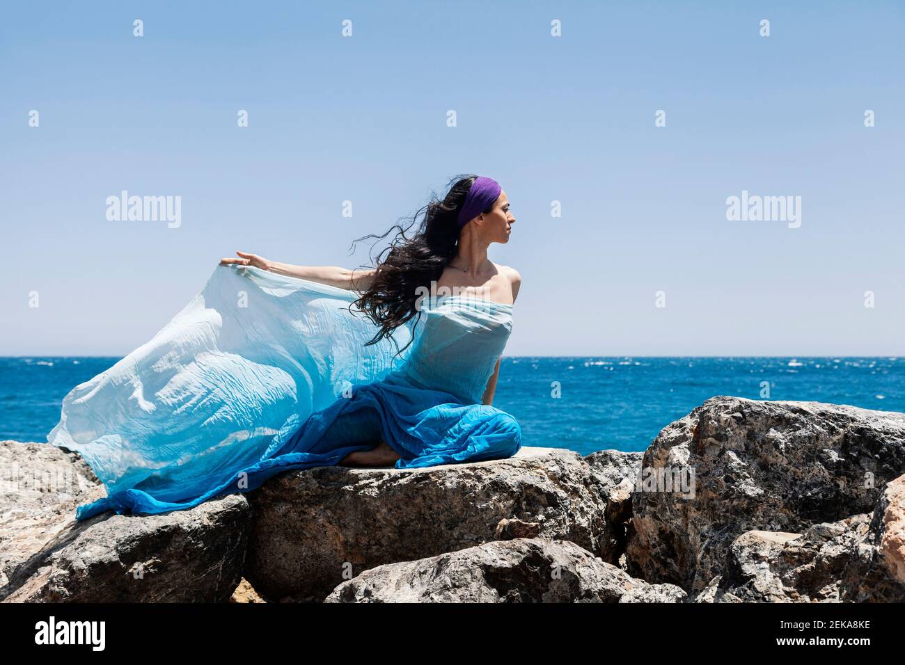 Ballerina femminile che tiene la sciarpa mentre si siede sulla roccia dal seashore durante il giorno di sole Foto Stock