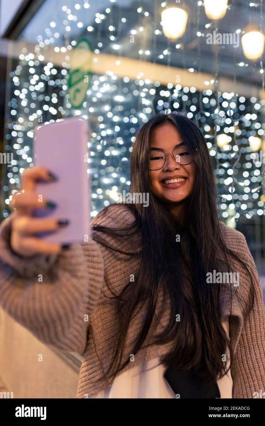 Giovane donna allegra che prende selfie attraverso smartphone contro illuminato parete di vetro Foto Stock