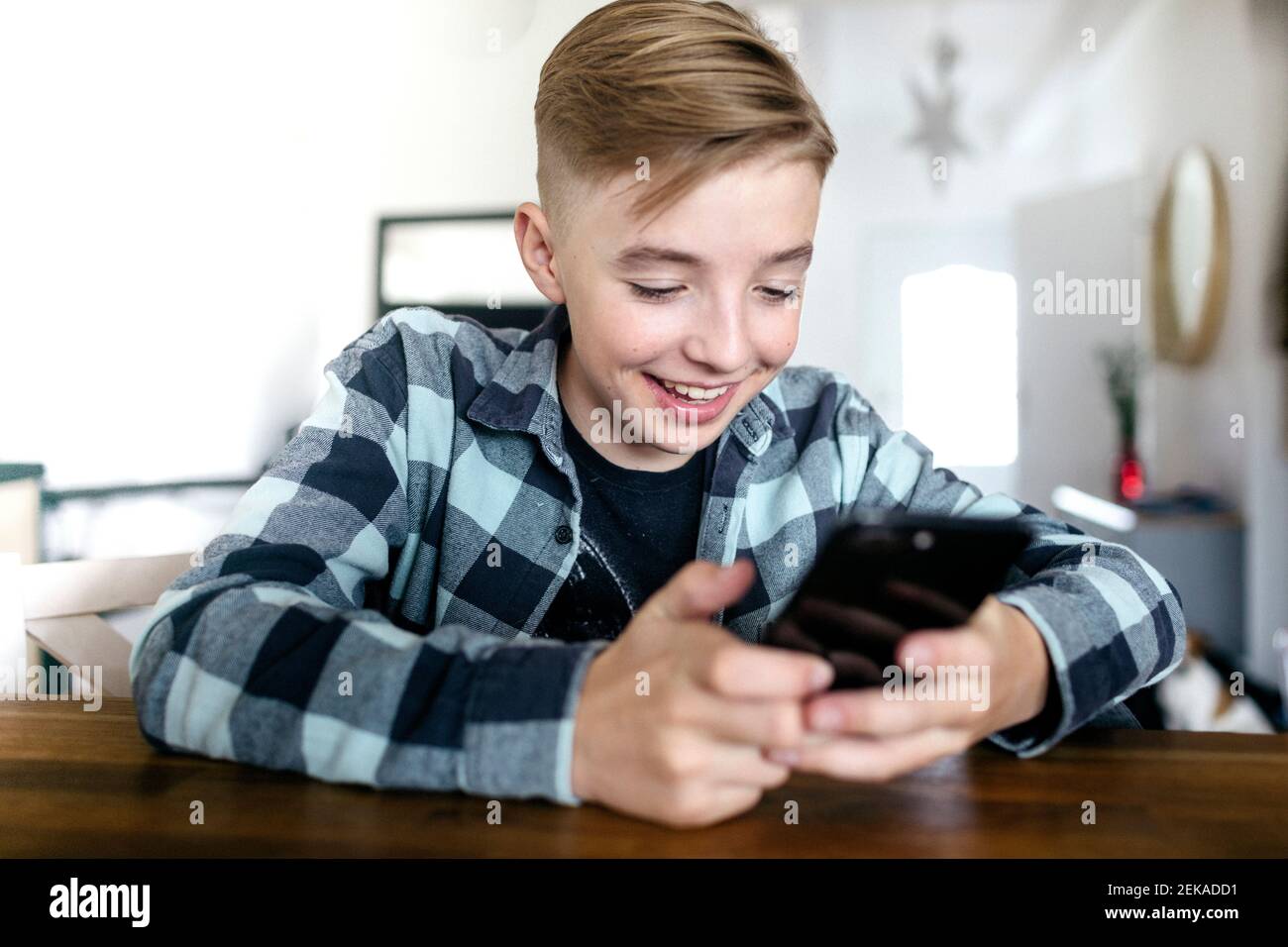 Ragazzo sorridente usando lo smartphone mentre si siede al tavolo in salotto Foto Stock