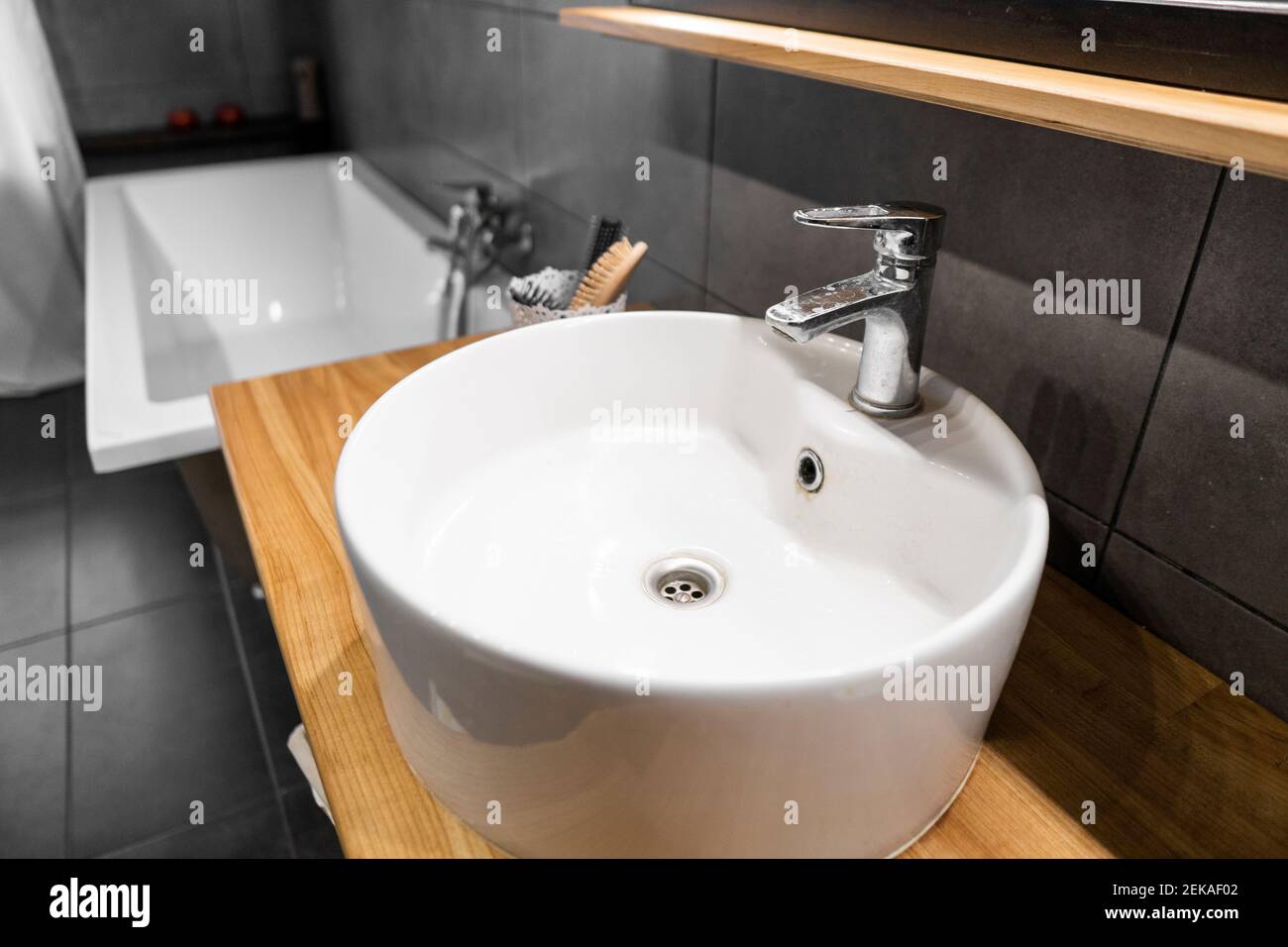 Interno del bagno con piastrelle grigie su una parete, elegante lavello rotondo e rubinetto su un piano di lavoro in legno di quercia. Minimalismo all'interno del bagno. Foto Stock