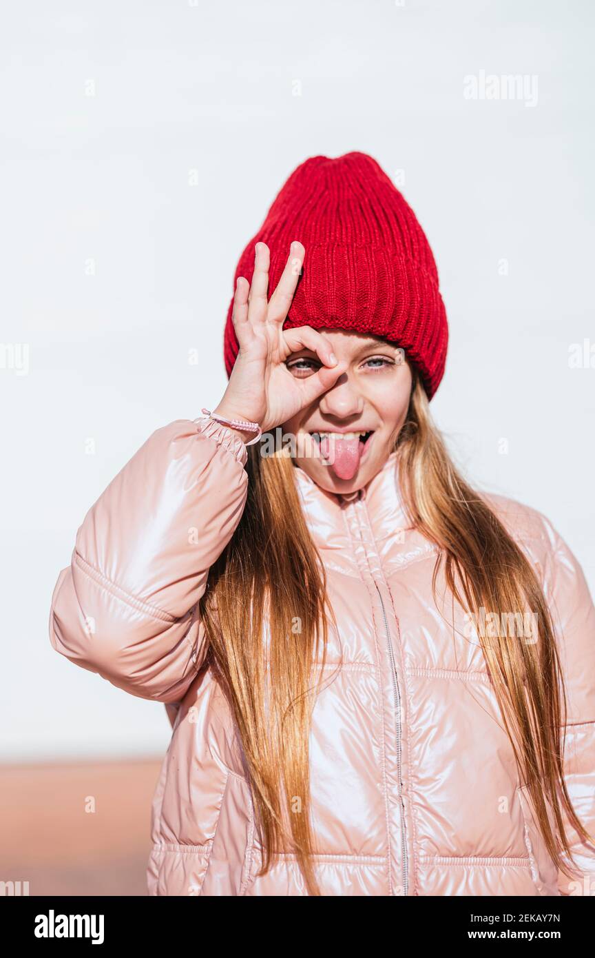 Giocosa ragazza che si stacca la lingua mentre gesturing OK segno contro parete Foto Stock