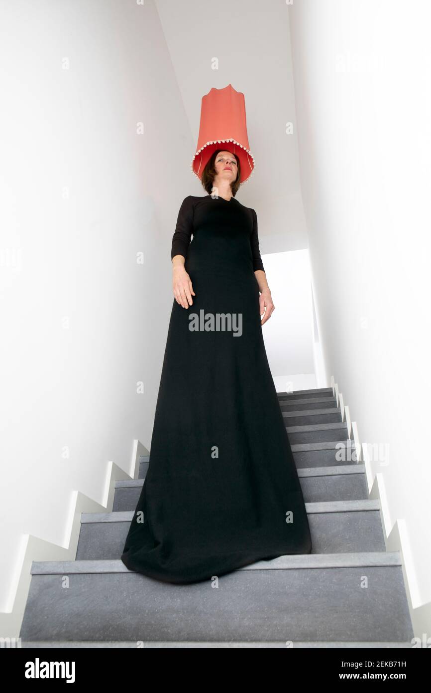 Donna in abito nero con paralume di lampada di pesca sulla testa in piedi su scala Foto Stock
