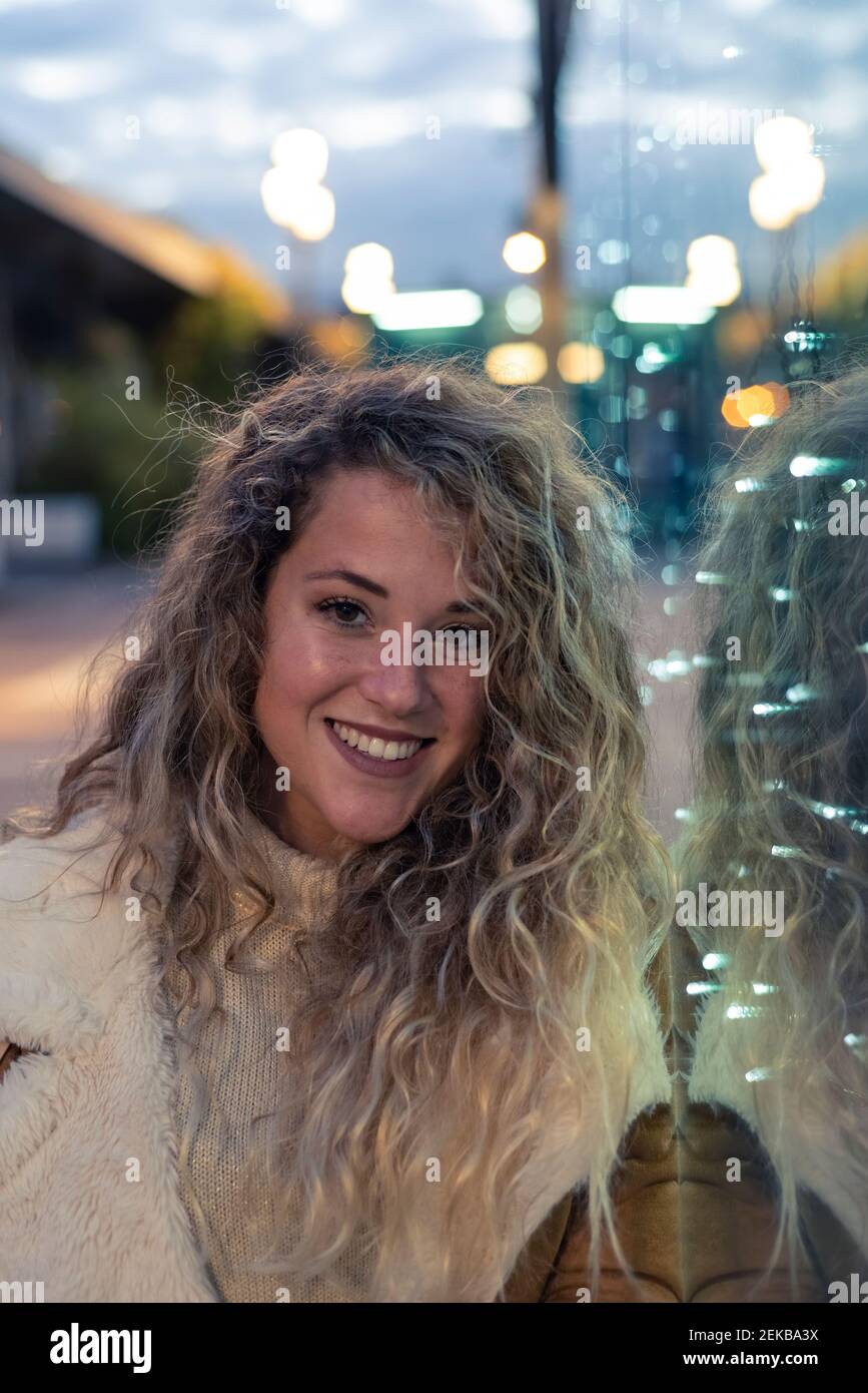 Giovane donna sorridente appoggiata su una parete di vetro illuminata al tramonto Foto Stock