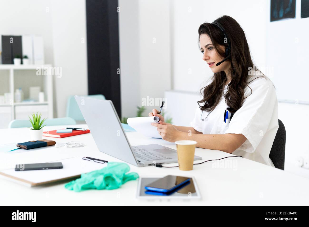 Giovane dottore femminile che scrive su blocco note mentre consultandosi in linea con computer portatile in clinica Foto Stock