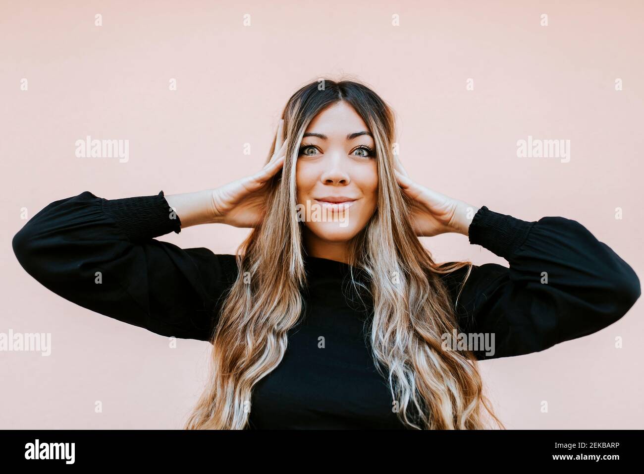 Sorridente giovane donna che tocca la testa mentre si alza contro il muro Foto Stock