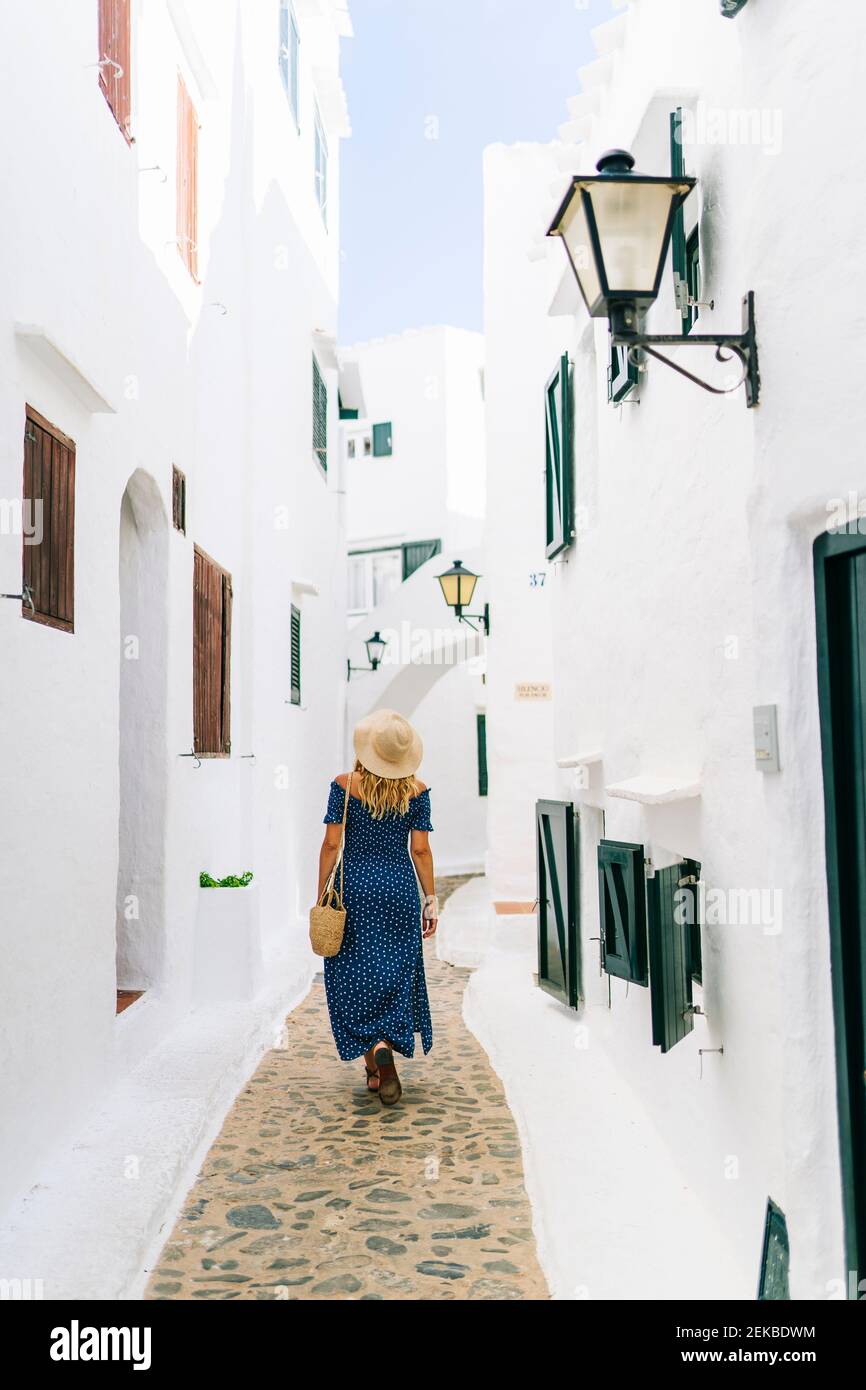 Donna matura che indossa cappello a piedi in vicolo tra le case a Binibeca villaggio, Minorca, Spagna Foto Stock