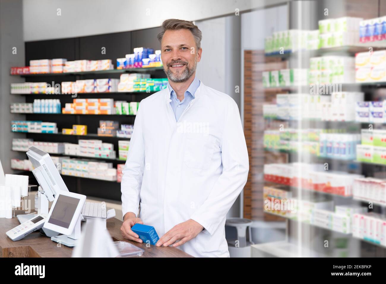 Sorridente farmacista maturo con la scatola della medicina in piedi al checkout della farmacia Foto Stock