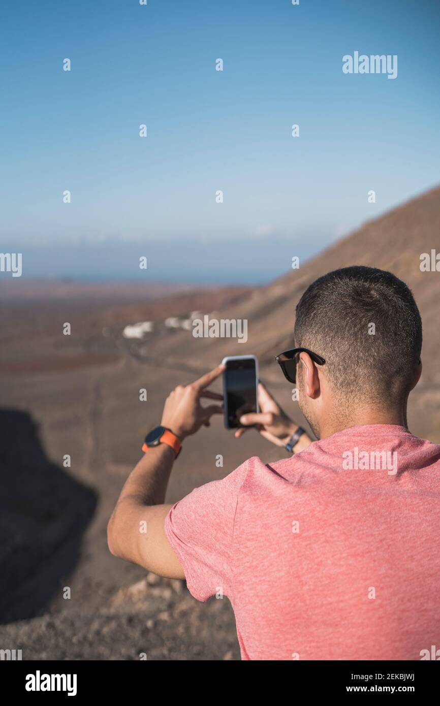 Giovane turista maschile fotografando attraverso lo smartphone al punto di osservazione Femés, Lanzarote in giornata di sole, Spagna Foto Stock
