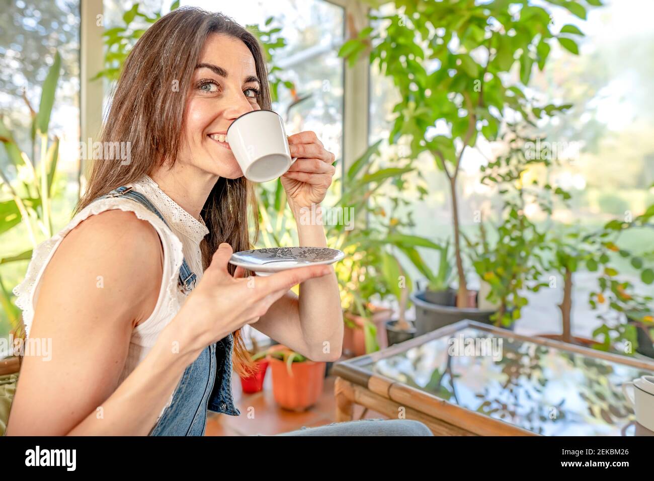 Donna sorridente con gli occhi grigi che beve caffè in sala da sole Foto Stock