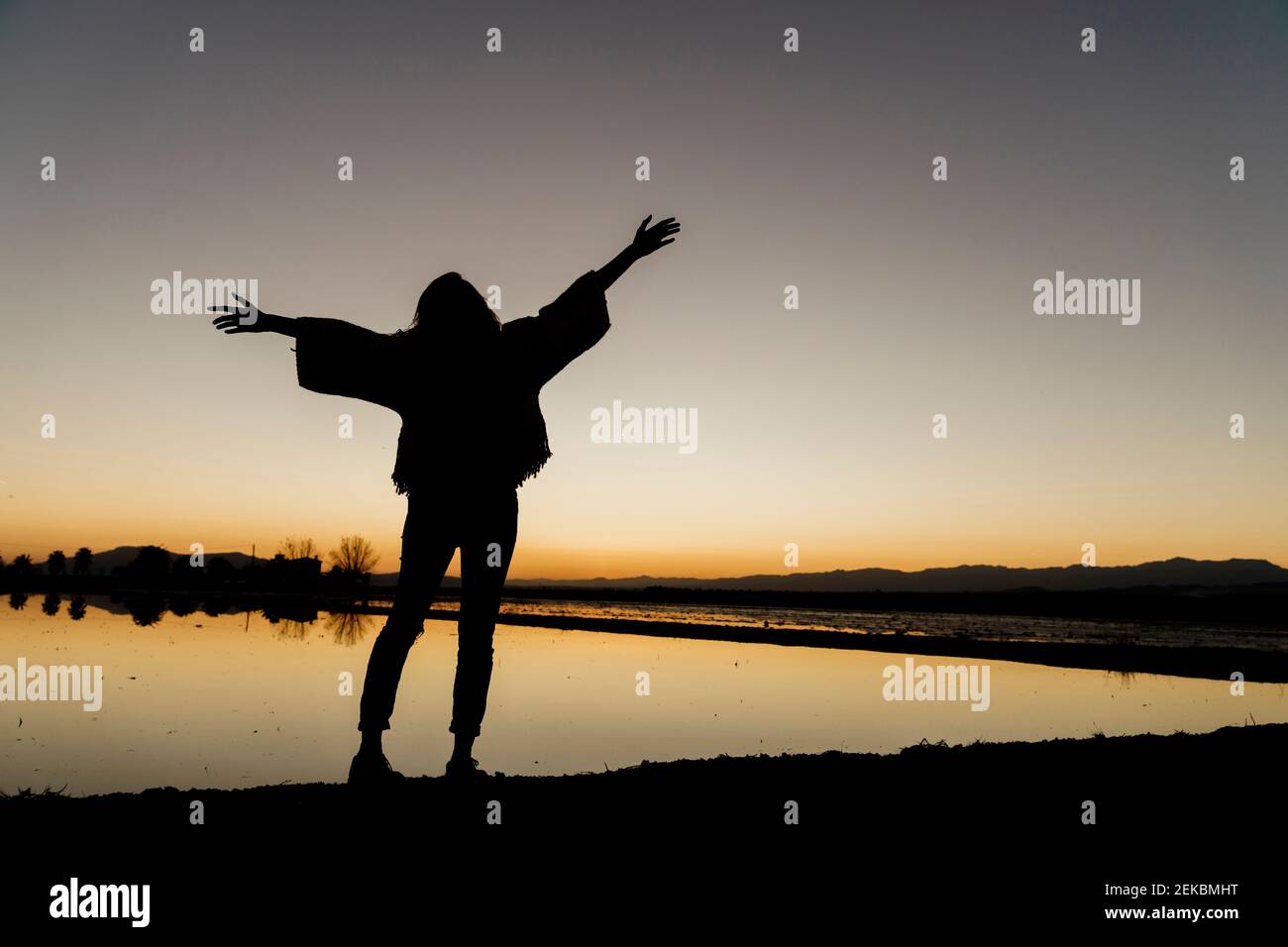 Giovane donna con le braccia stese in piedi dal fiume durante il tramonto a Ebro delta, Spagna Foto Stock