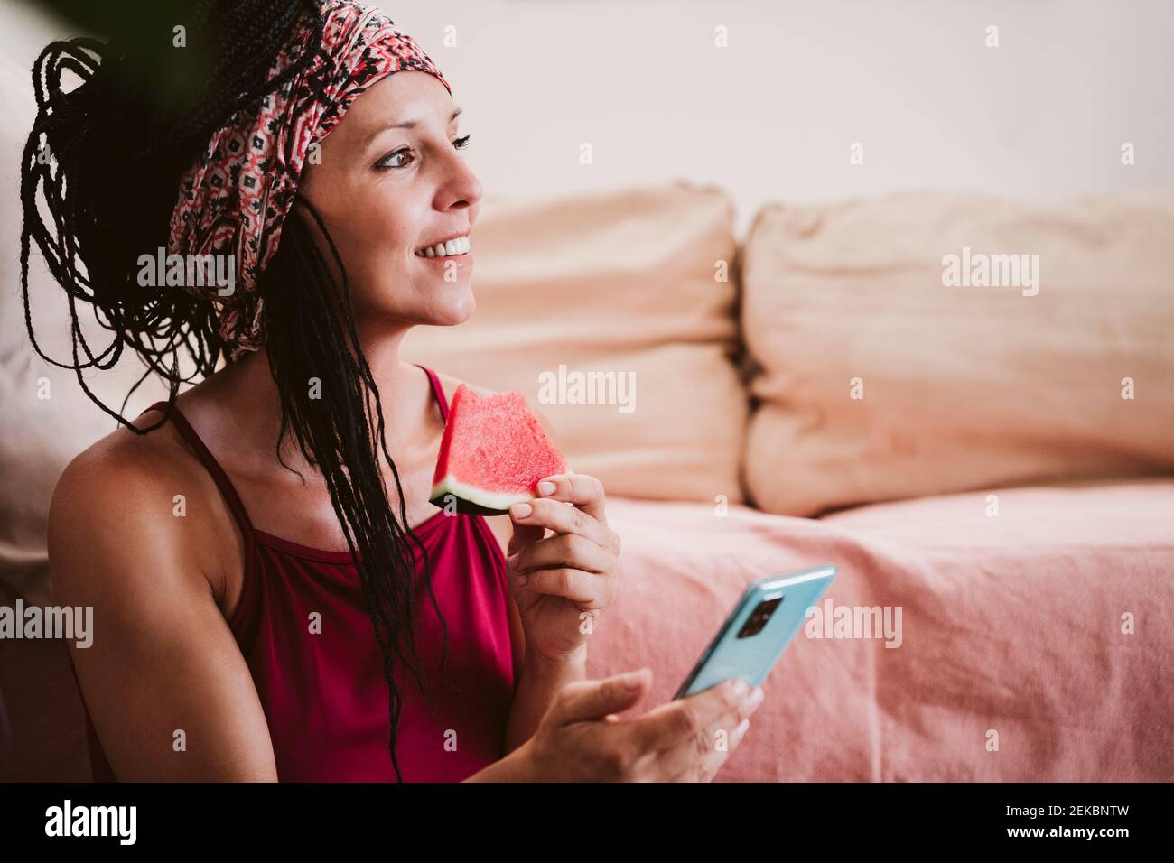 Donna con telefono cellulare che mangia l'anguria mentre si siede a casa Foto Stock