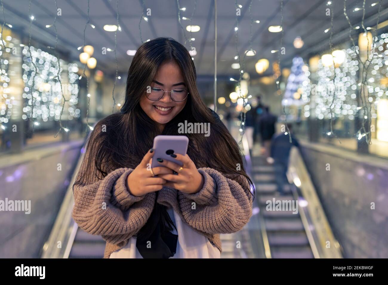 Giovane donna che usa lo smartphone mentre si sta in piedi contro il vetro illuminato muro nel centro commerciale Foto Stock