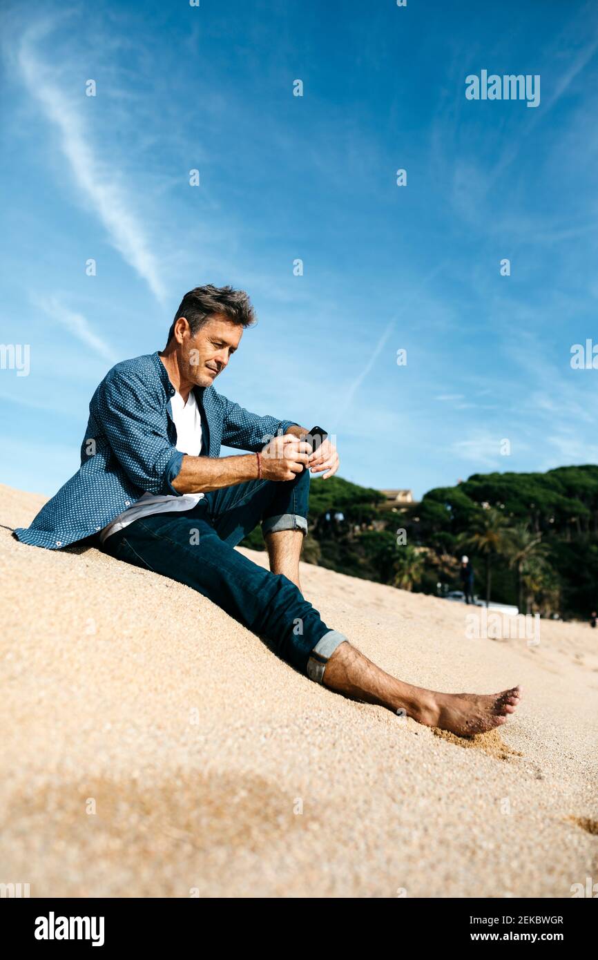Uomo che usa il telefono seduto in spiaggia contro il cielo Foto Stock