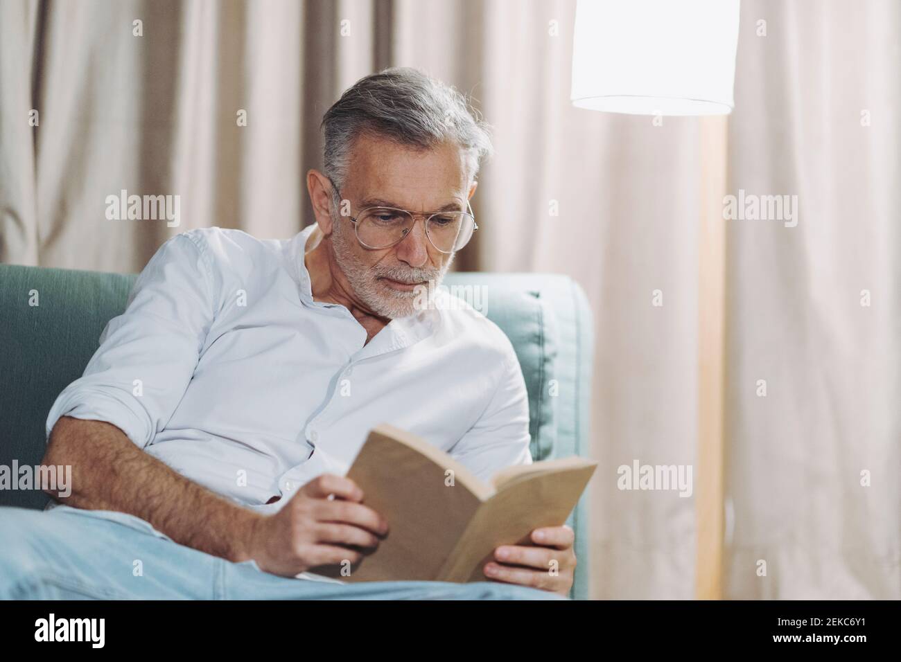 Un uomo curioso che legge libro mentre si siede sul divano dell'hotel camera Foto Stock