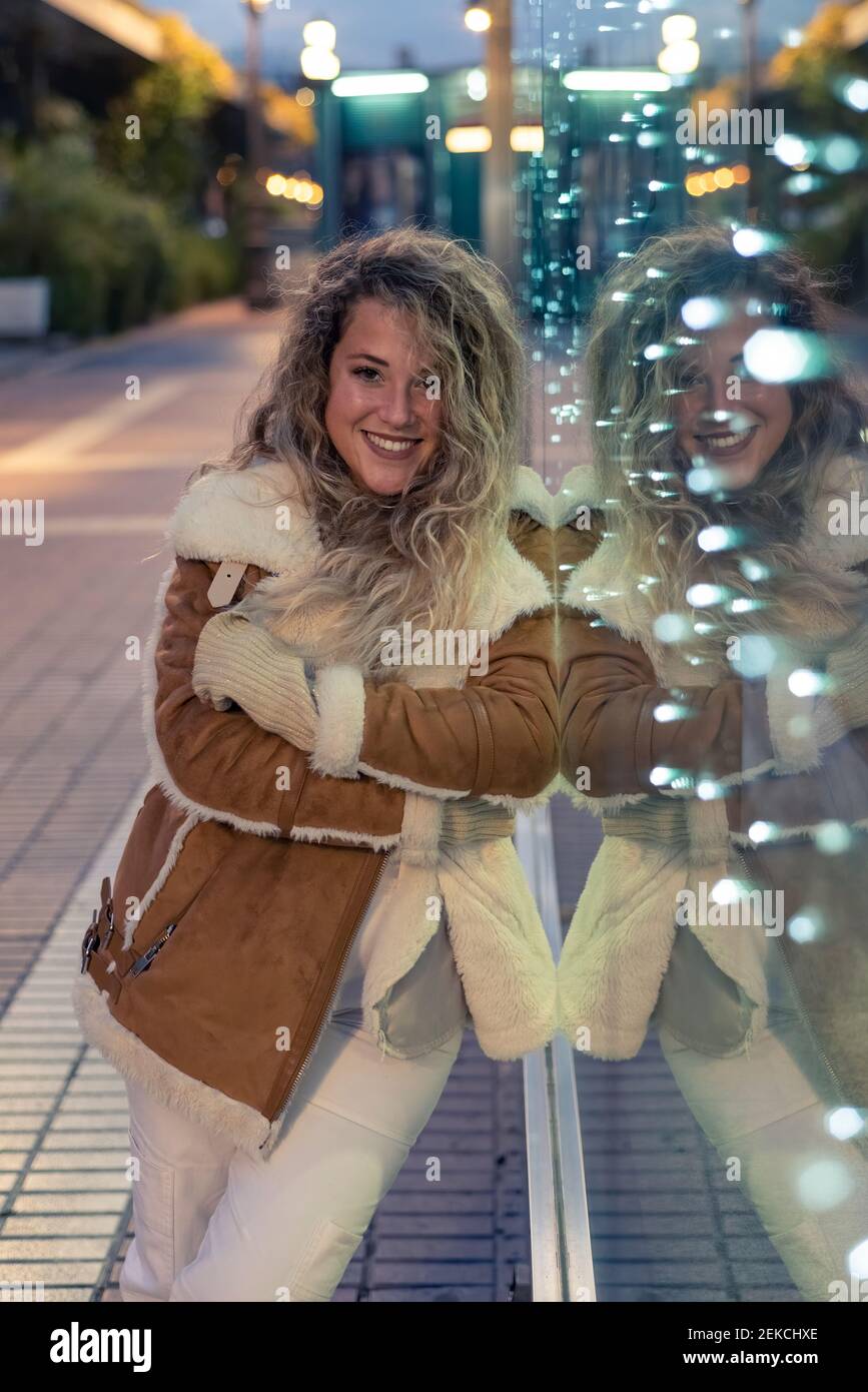 Donna sorridente con le braccia incrociate appoggiate su una parete di vetro illuminata al crepuscolo Foto Stock