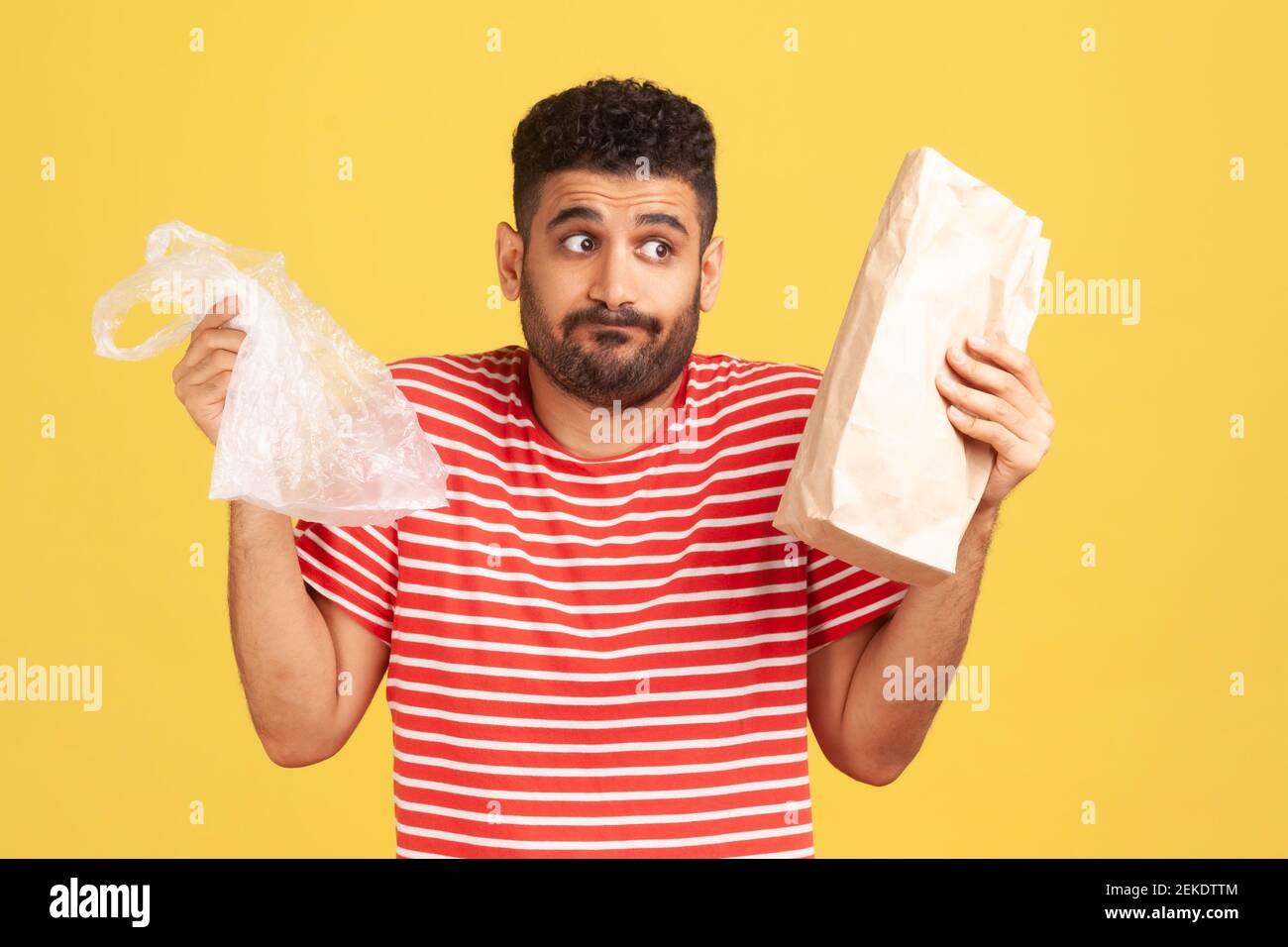 L'uomo confuso e perplesso con la barba che tiene in borse di plastica e di carta, cercando di fare la scelta giusta, riducendo il consumo di plastica. Riprese in studio al coperto Foto Stock