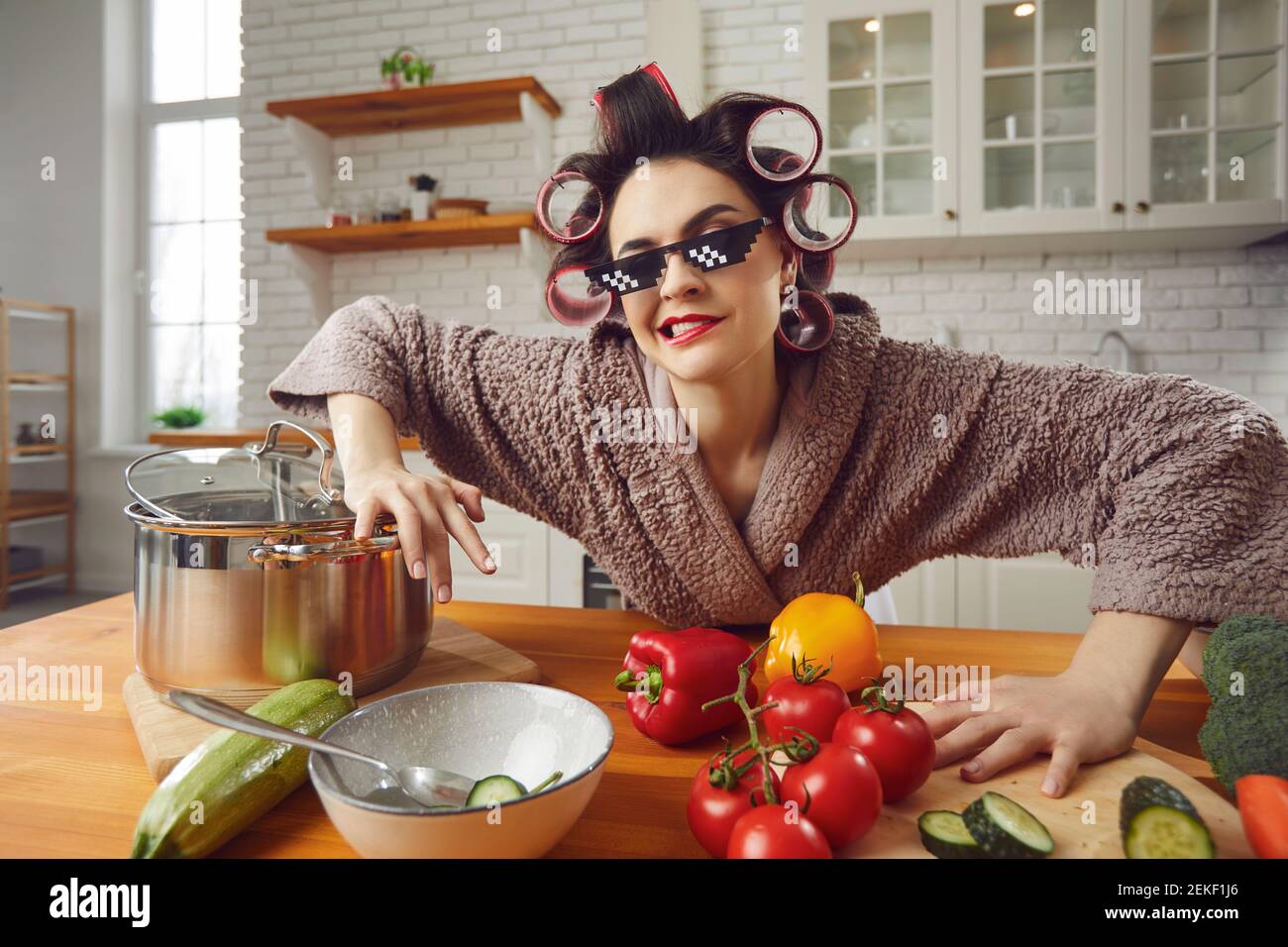 Divertente e bella donna pazza in arricciacapelli e accappatoio preparando la colazione in cucina. Foto Stock