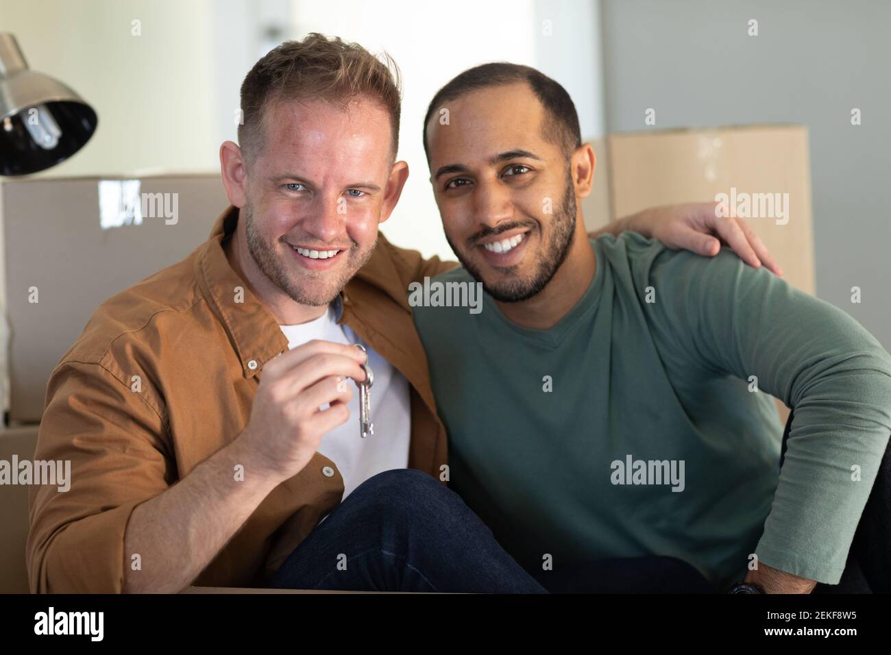 Multi etnico gay maschio coppia seduta circondata da scatole che guardano alla fotocamera sorridendo a casa Foto Stock