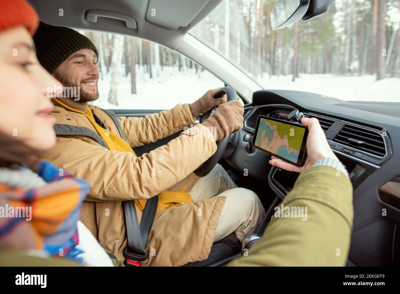 Felice giovane uomo attivo in winterwear caldo guardando la mappa sullo schermo dello smartphone tenuto dalla moglie durante la guida durante i viaggi invernali Foto Stock