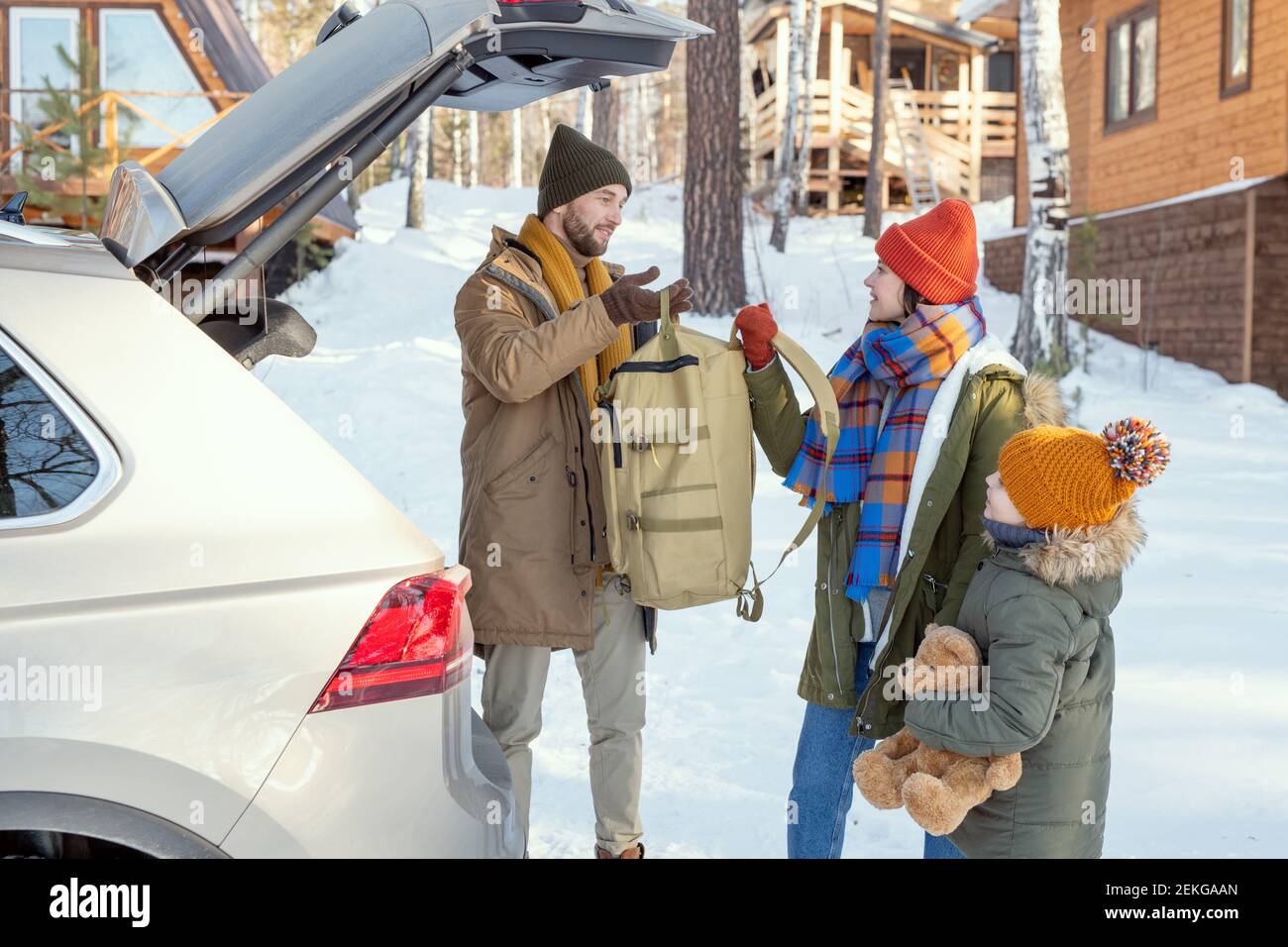 Giovane famiglia di tre in winterwear caldo che prende il loro buggage fuori dal vano bagagli mentre ci si trova sul retro dell'auto durante i viaggi invernali Foto Stock