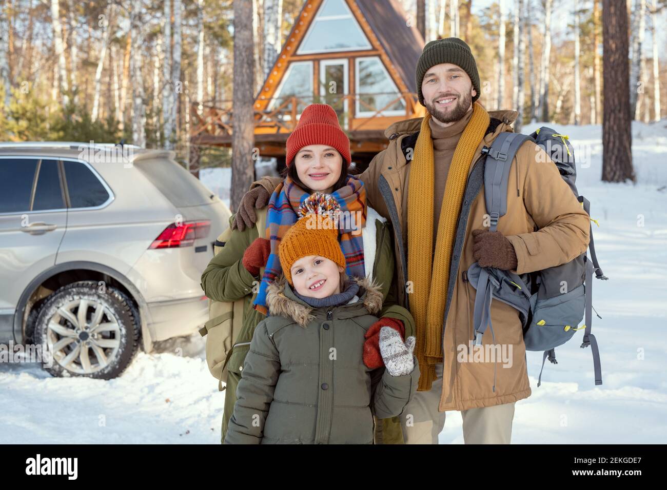 Felice giovane famiglia di tre in caldo Winterwear in piedi davanti alla telecamera contro la casa di campagna e guardando voi fuori Foto Stock