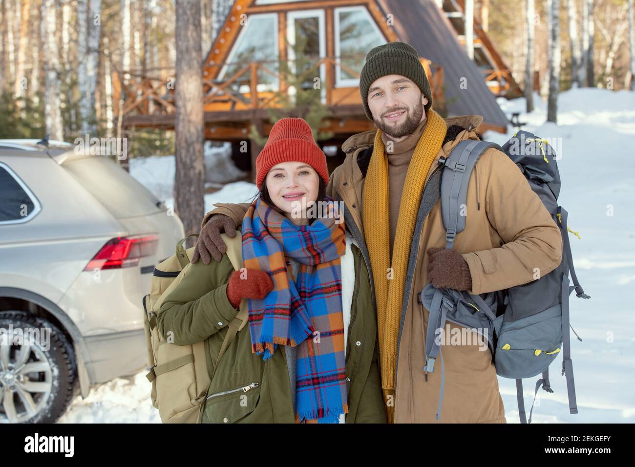 Giovane coppia affettuosa in winterwear caldo in piedi davanti a. fotocamera contro la loro auto e casa di campagna e guardando voi fuori Foto Stock