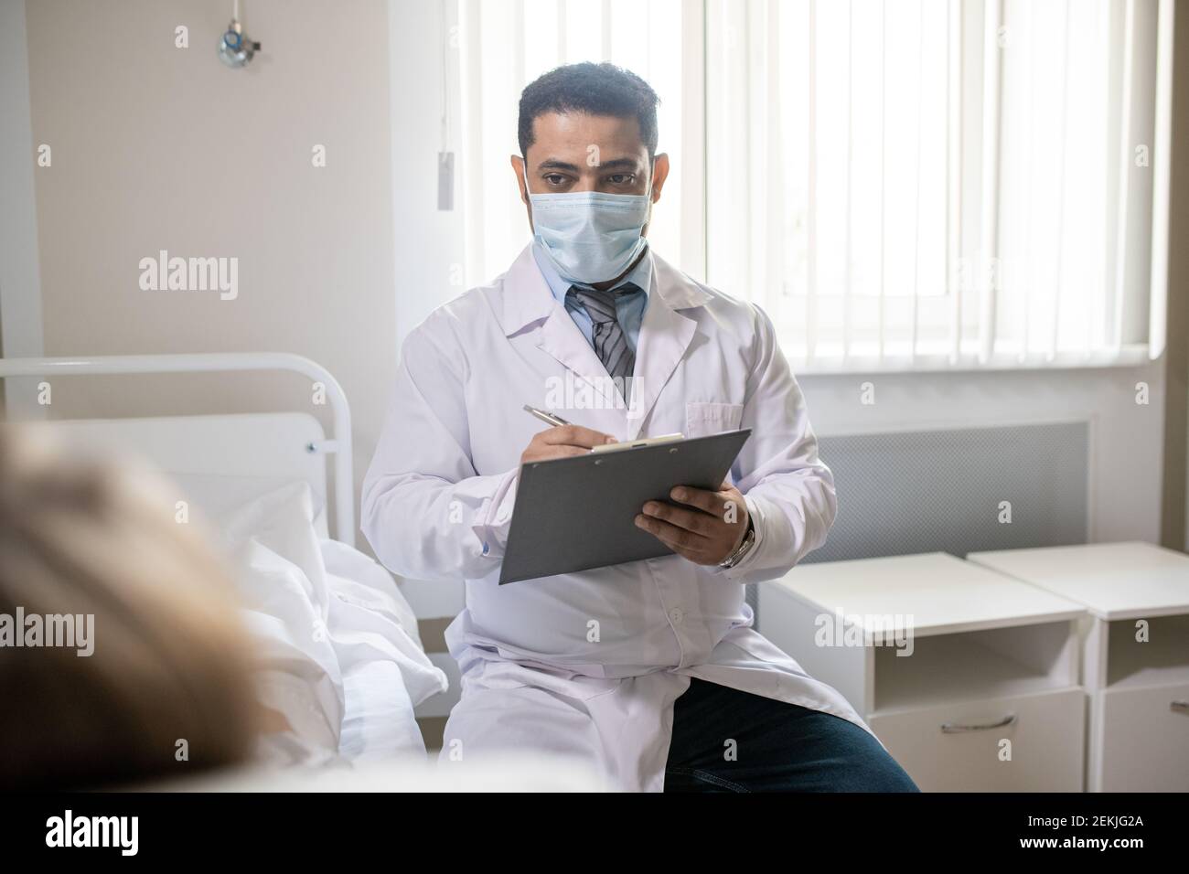 Giovane medico in whitecoat e maschera protettiva scrivere medico dati nel documento e esame del paziente durante la conversazione Foto Stock