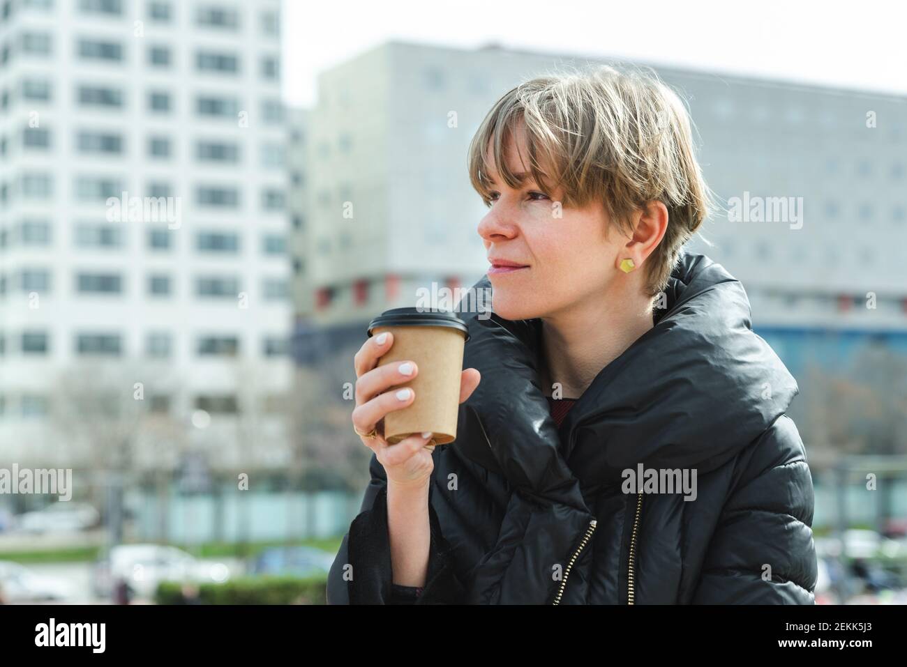 bionda giovane donna vestendo abiti invernali e tenendo una tazza di carta di caffè take away con uffici edifici sullo sfondo. Copia spazio a sinistra Foto Stock