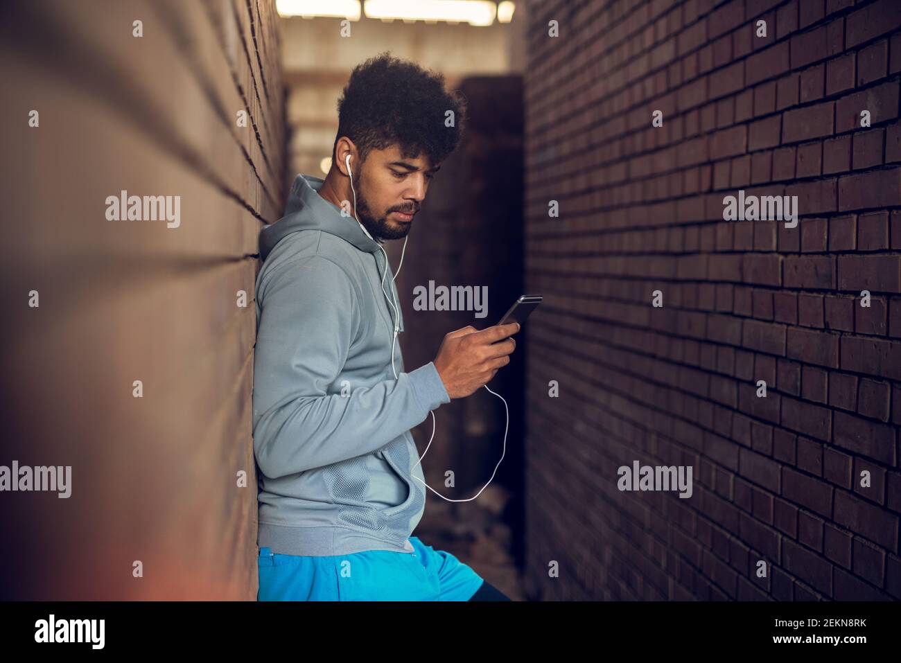 Giovane uomo afro-americano con cuscinetti appoggiati alla parete che ascolta la musica e tiene un cellulare tra due pareti esterne. Foto Stock