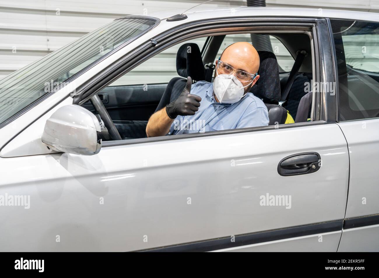 Un tecnico in un'ispezione periodica di un veicolo che mostra i pollici all'interno di un'auto, protetto da maschera facciale, guanti e occhiali Foto Stock