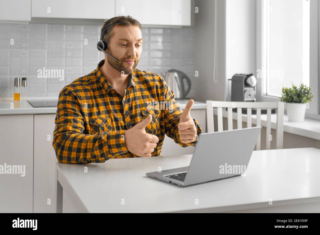 Un uomo durante una videochiamata online. Si siede a casa davanti al monitor di un computer portatile e mostra i pollici verso l'alto. Concetto di tecnologie moderne per la comunicazione Foto Stock