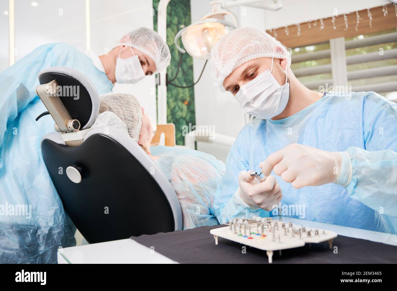 Dentista in primo piano, indossando abiti sterili monouso, posizionando l'ugello chirurgico sul trapano prima dell'installazione dell'impianto. Paziente e assistente su sfondo sfocato. Concetto di posizionamento dell'impianto. Foto Stock